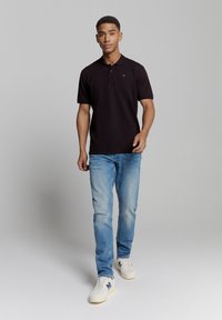 Zwarte poloshirt met een kraag, korte mouwen, gecombineerd met lichtblauwe jeans en witte sneakers. De polo heeft een klein logo op de borst.