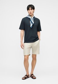 Fiatal férfi sötétkék pólóban, fehér-kék mintás sállal, bézs rövidnadrágban és sötétbarna szandálban, egyszínű háttér előtt.