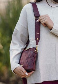 Burgundská kůže křížová taška s texturovanými detaily, zlatými prvky a nastavitelným popruhem, který ladí se světle šedým pleteným svetrem.