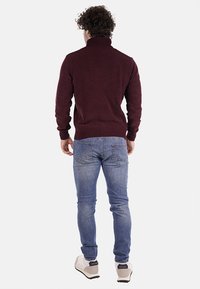 Pullover lavorato a maglia bordeaux con colletto alto, abbinato a jeans azzurri chiari. L'outfit presenta una finitura testurizzata e una vestibilità standard.