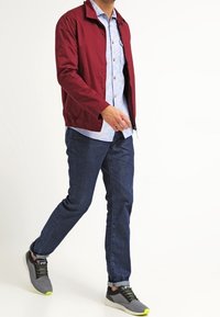 Mies, joka kävelee punaisessa takissa, vaaleansinisessä paidassa, tummansinisissä farkuissa ja harmaissa lenkkareissa, joissa on neonkeltaisia yksityiskohtia.