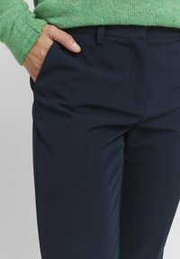 Granatowe spodnie o gładkiej fakturze, z przednimi kieszeniami i dopasowanym krojem, w połączeniu z zielonym sweterkiem o fakturze.