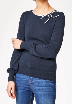 Kvinde iført en mørkeblå, langærmet sweater med hvide kanter og en stribet sløjfe nær udskæringen, kombineret med blå jeans.