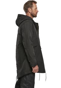 Chaqueta negra con capucha de corte largo, con cierre frontal de cremallera, dos bolsillos laterales y cordones ajustables en el dobladillo, hecha de un tejido texturizado.