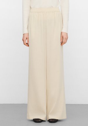 Creme kleurige wijde broek, gedragen met een licht shirt met lange mouwen, van taille tot voeten getoond tegen een effen achtergrond.