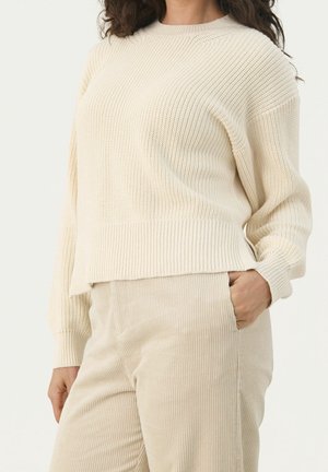 Woman wearing a cream-colored ribbed knit sweater and beige corduroy pants with one hand in pocket, shown from neck to hips.