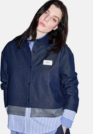 Joven con cabello largo y oscuro que lleva una camisa azul a rayas de varias capas y una chaqueta corta de mezclilla oscura, con las manos en los bolsillos y la cabeza ligeramente inclinada.