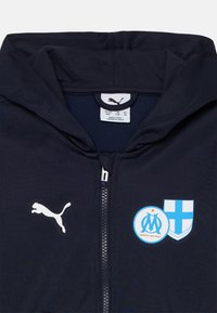 Jūros mišinio džinsinis su užtrauktuku, su baltu Puma logotipu ir Marselio herbu. Minkšta medžiaga su lygiu paviršiumi ir reguliuojama gaubtu.