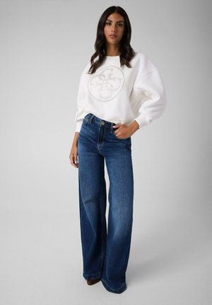 Femme aux cheveux foncés portant un sweat-shirt blanc avec un logo circulaire et un jean bleu à jambes larges, debout devant un fond gris uni.