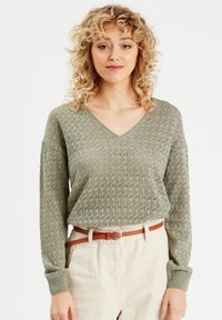 Maglione lavorato a maglia verde oliva con scollatura a V, maniche lunghe e motivo testurizzato. Abbinato a pantaloni crema e una cintura marrone.