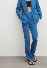 Blue tailored blazer and matching trousers made from a smooth fabric, with large pockets and a high-waisted fit, paired with black flip-flops.