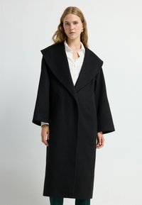 Lange zwarte jas met een brede kraag, gestructureerd ontwerp en klokvormige mouwen. Bevat zijzakken en een strakke stikselafwerking.