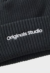 Gorro de punto negro acanalado con puño doblado. Presenta una etiqueta blanca que dice "Originals Studio" en la parte delantera. Tejido texturizado.