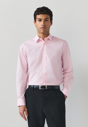 Homme portant une chemise rose clair à manches longues boutonnée, rentrée dans un pantalon noir avec une ceinture noire, debout devant un fond clair uni.