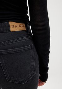 Črne jeans hlače iz denima z rjavim usnjenim označevalcem, na katerem piše "NA-KD." Značilnosti vključujejo srednje višji pas in zasnovo petih žepov z vidnim šivanjem.