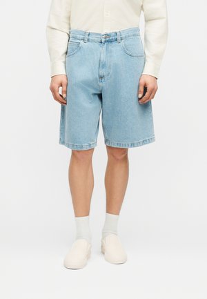 Osoba ubrana w jasnoniebieskie dżinsowe szorty, białą koszulę z długim rękawem, białe skarpetki i kremowe buty wsuwane, stojąca na białym tle.