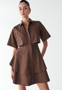 Rjava, kratkodlaka obleka s kratkimi rokavi, ovratnikom, zasnovo s preklopom, plastenim krilom in gladko teksturo. Ima ravno prileganje in minimalističen slog.