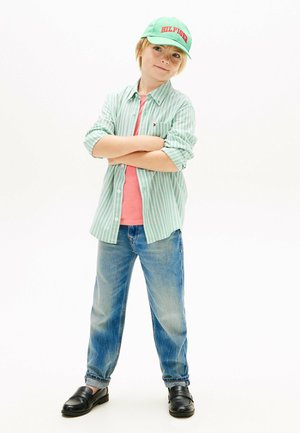 Ung dreng iført grønstribet skjorte, lyserød t-shirt, blå jeans, sorte sko og grøn kasket med teksten "HILFIGER", stående med armene over kors.