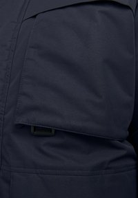 Marineblauwe jas met een grote klepzak, een gladde textuur en duurzame stof met een klein verstelbaar riempje.