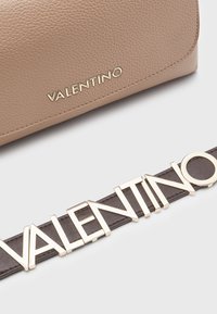 Brun läderhandväska med strukturerad yta, prydd med en guldig "Valentino"-logotyp. En brun bälte med metallisk guldtext är placerad under.