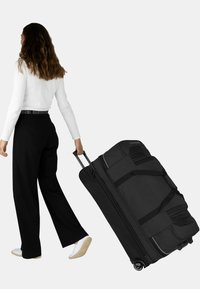 Schwarze Rollreisetasche mit mehreren Taschen, Stoffstruktur und verstärkten Griffen. Person zieht die Tasche und trägt schwarze Hosen und ein weißes Oberteil.