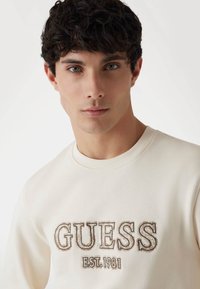 Felpa color crema con ricamo testurizzato "GUESS EST.1981" in marrone scuro. Design classico con collo a girocollo e finitura in tessuto morbido.