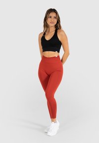 Top corto a righe nere abbinato a leggings arancioni a vita alta. La modella indossa sneakers bianche su uno sfondo neutro.