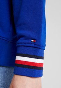 Modrá mikina s texturovanou látkou, s žebrovanými manžetami zdobenými červenými, bílými a námořnickými pruhy. Na manžetě je viditelná logová značka.