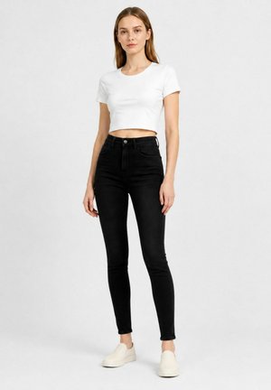 Giovane donna in piedi che indossa una t-shirt bianca corta, jeans skinny neri a vita alta e sneakers bianche slip-on su uno sfondo semplice.