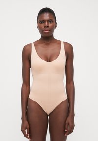 Nude heldrakt med V-hals, brede skulderstropper og sømløst design, laget av elastisk stoff. Glatt tekstur, minimal detalj.