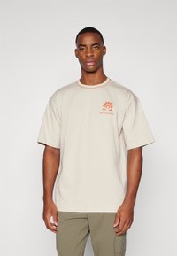 Camiseta de algodón beige de manga corta, con costuras de acento en naranja y un logo que presenta un engranaje y un ojo en la parte izquierda del pecho.