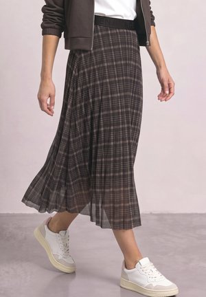 Personne portant une jupe plissée à carreaux marron, des baskets blanches avec des semelles beiges, un haut blanc et une veste marron avec les manches retroussées.