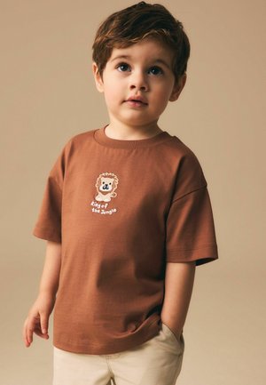 Mladý chlapec s hnědými vlasy, oblečený v hnědém tričku s lvem a nápisem "Král džungle", béžové kalhoty, dívá se mírně vzhůru.