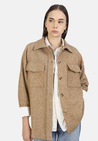 Bruine jasje met een visgraatpatroon, voorzien van twee borstzakken, een grote kraag en een knoopsluiting, gedragen over een lichtgekleurde blouse.