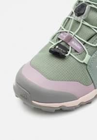 Zaļi un pelēki pārgājienu apavi ar strukturētu augšdaļu, rozā akcentiem, GORE-TEX zīmolu un gumiju saistēšanas sistēmu.