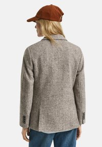 Blazer grigio in lana con una finitura texturizzata, forma su misura, colletto a punte e spacco singolo sul retro. Abbinato a un berretto in corda color ruggine.