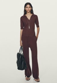 Bordeaux strikbluse med korte ærmer og v-udskæring, parret med matchende bukser. En sort håndtaske og sandaler fuldender outfitet.