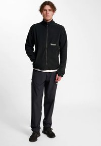 Zwarte fleecejas met volledige ritssluiting, hoge kraag en logodetail. Gecombineerd met marineblauwe broek en zwarte schoenen, staand op een neutrale achtergrond.