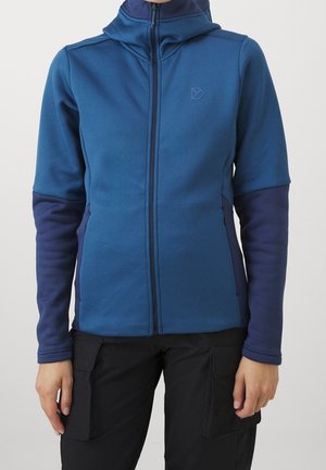 Person wearing blue zip-up jacket with darker blue sleeves and black cargo pants against a plain light background.