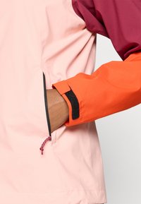 Veste imperméable avec un corps rose, des manches bordeaux et des poignets orange. Elle comporte une patte Velcro et un cordon de serrage ajustable à l'ourlet.