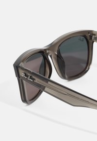 Ochelari de soare Ray-Ban cu un cadru translucid de culoare gri închis, lentile tonificate în verde și maro și un logo argintiu pe braț.