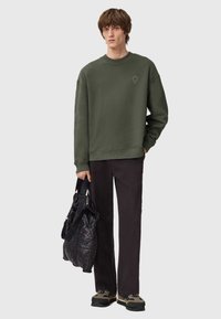 Felpa verde oliva con scollo rotondo, spalle cadenti; abbinata a pantaloni neri e una borsa nera trapuntata; modello in atteggiamento casual.