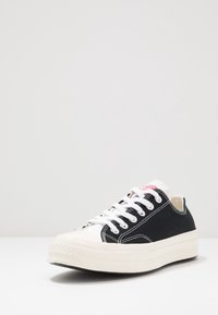 Niskie czarno-białe sportowe buty z białymi sznurowadłami, grubą białą podeszwą i czerwonym logo na języku, prezentowane na białym tle.