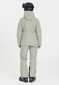 Persona che indossa un completo da sci grigio chiaro, un casco nero e scarponi da sci, di spalle su sfondo bianco.