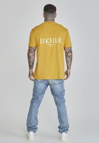 Žuta majica s kratkim rukavima s bijelim natpisom "SIKSILK" na leđima, nosi se s svijetloplavim trapericama i bijelim tenisicama.