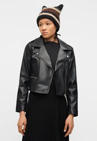 Νέα γυναίκα που φορά μαύρο δερμάτινο μπουφάν μοτοσικλετιστή, μαύρο φόρεμα και ριγέ πλεκτό σκουφάκι, στέκεται μπροστά σε ουδέτερο γκρι φόντο.