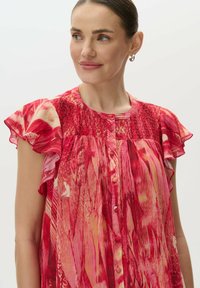 Rote gemusterte Bluse mit kurzen Rüschensleeves, die einen strukturierten Oberteil und eine Knopfleiste hat. Der Stoff zeigt abstrakte, lebendige Muster.