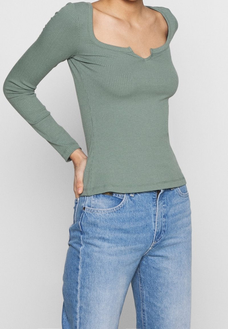 Langærmet, ribbet top i dæmpet grøn med firkantet halsudskæring og subtile udskæringsdetaljer, parret med lyseblå jeans.