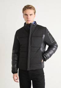 Black puffer jacket with a high collar, featuring crosshatch stitching, two front pockets, and a textured, shiny sleeve accent.