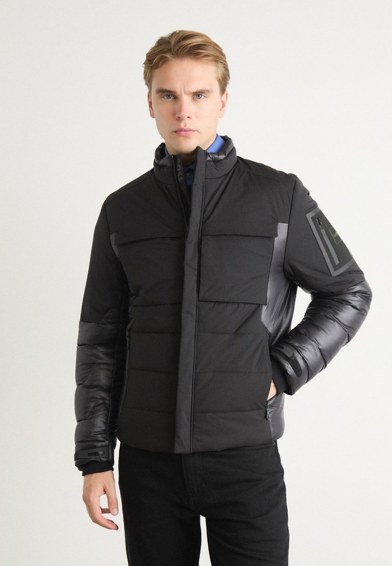 Black puffer jacket with a high collar, featuring crosshatch stitching, two front pockets, and a textured, shiny sleeve accent.